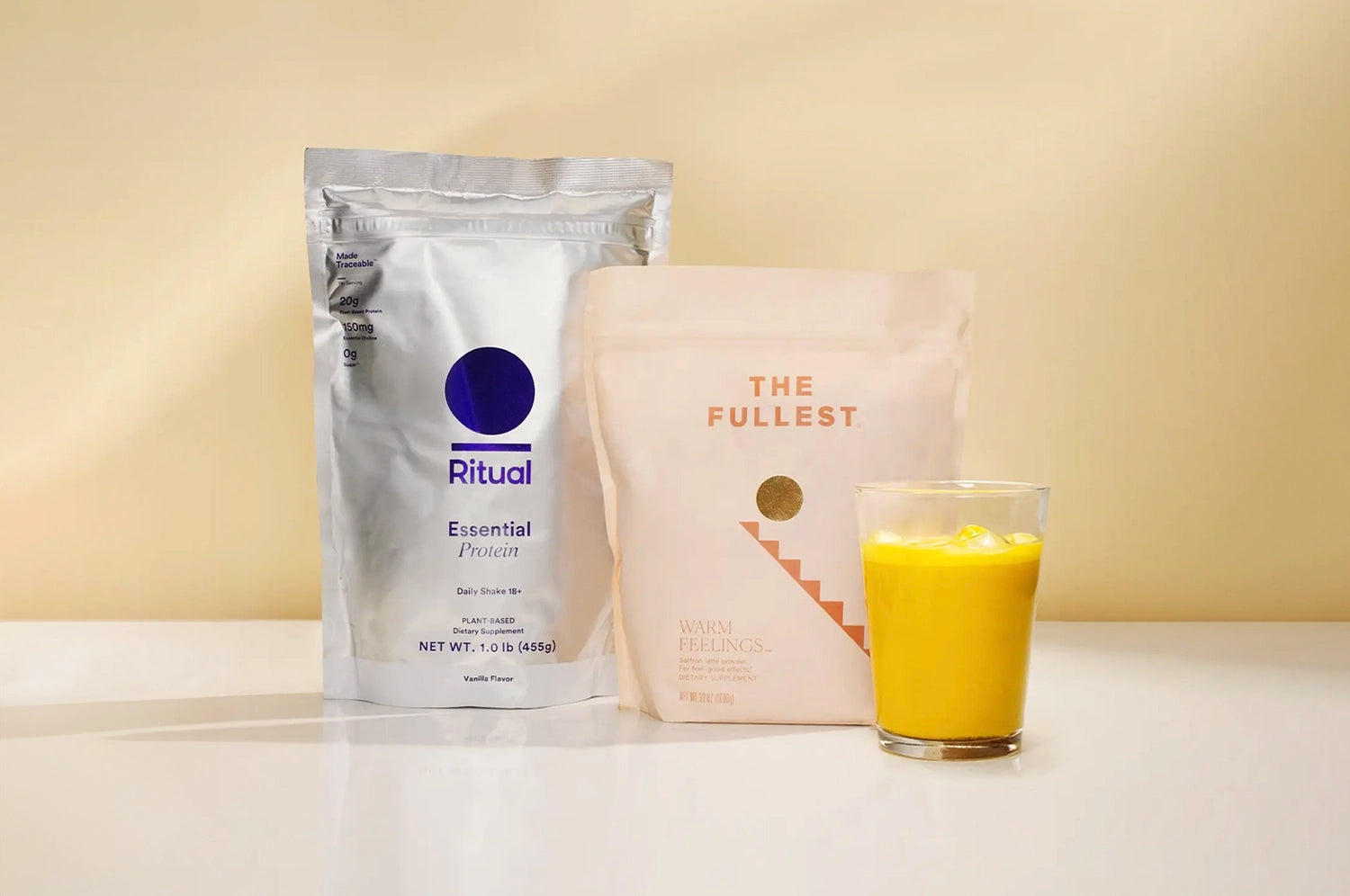 An image of The Fullest Warmest Feelings bag and a bag of Rituals Essential Protein Powder against an orange-blush toned background, with a saffron protein smoothie in a clear glass in the foreground.