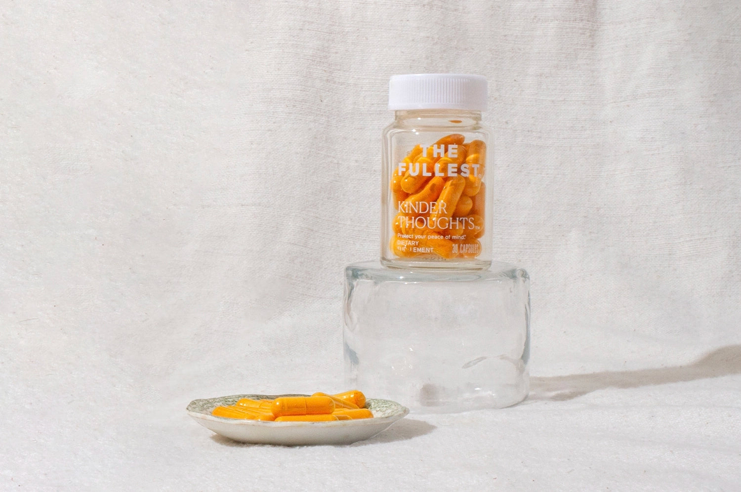 A clear pill bottle labeled "The Fullest Kinder Thoughts" filled with orange saffron and turmeric capsules, placed on a glass surface with additional capsules on a small plate nearby, against a textured white background.