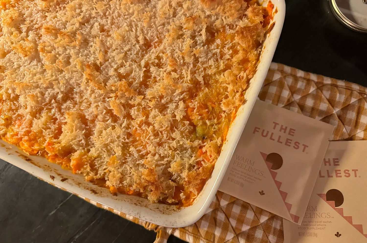 A golden, crispy-topped sweet potato casserole sits on a checkered cloth, paired with two sachets of THE FULLEST’s Warm Feelings saffron latte, adding a cozy, unique touch.