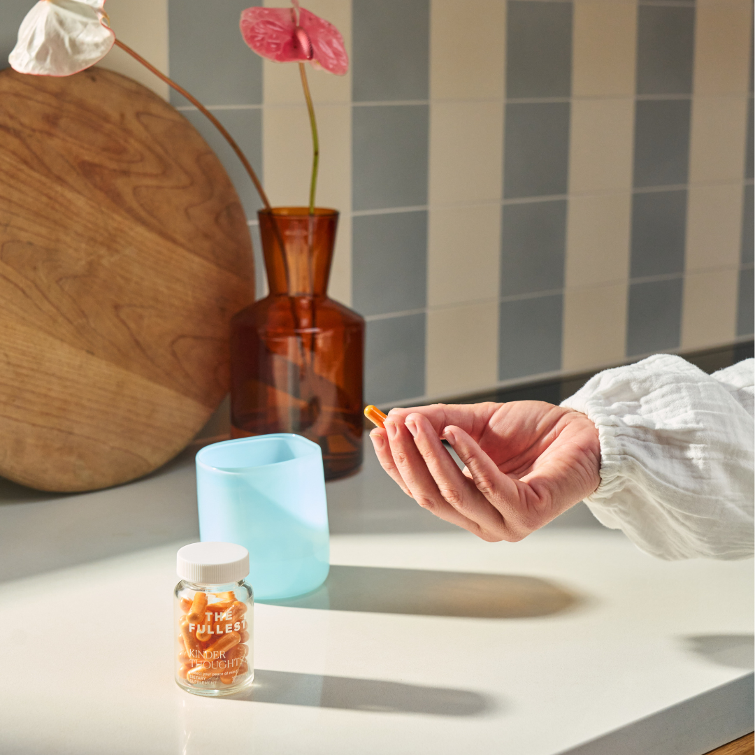 A person holds two Saffron Capsules from THE FULLEST above a kitchen counter with a blue cup, a glass bottle labeled "THE FULLEST," and a wooden cutting board in the background.
