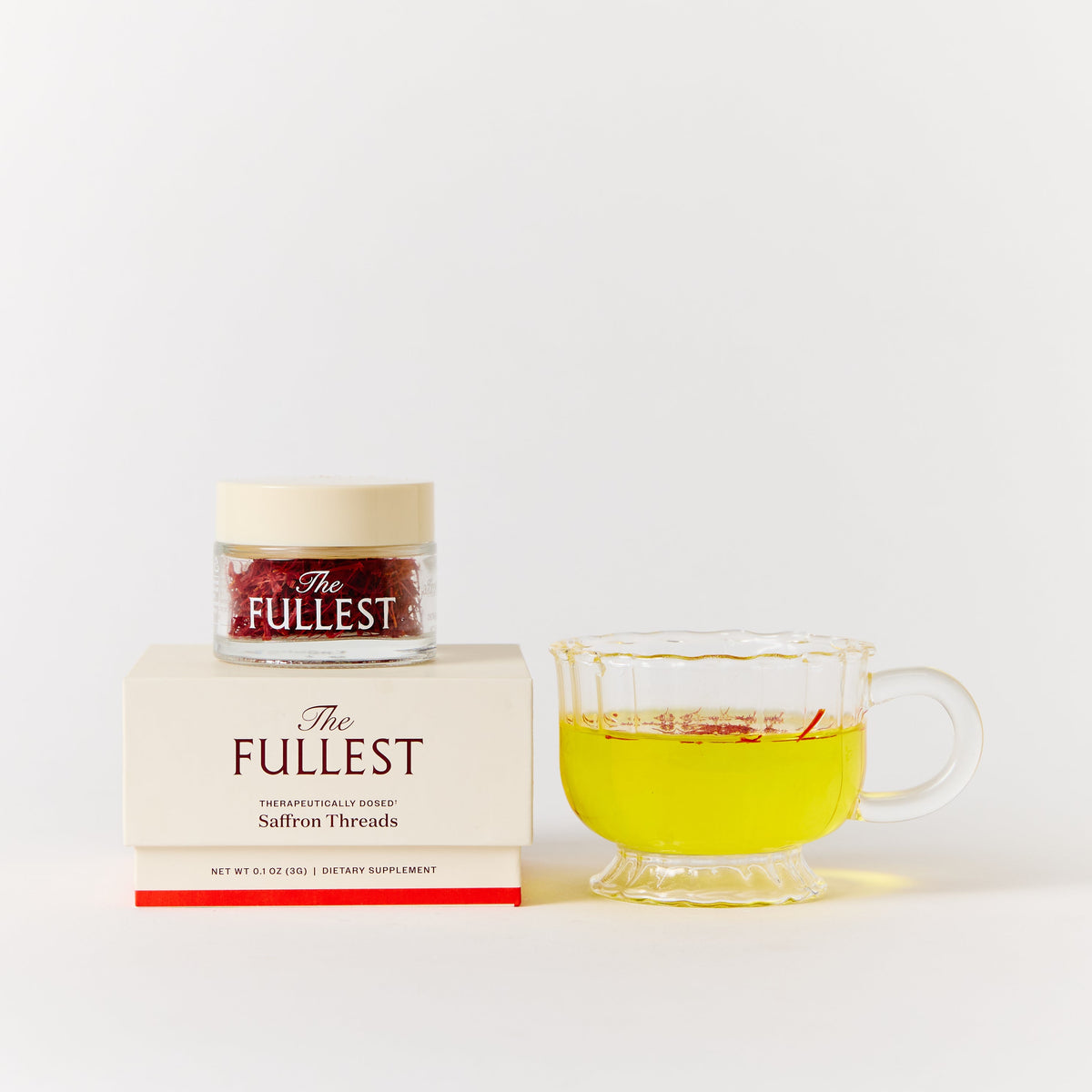 A glass jar of THE FULLEST Saffron Threads sits atop a square product box next to a glass cup of yellow saffron tea.
