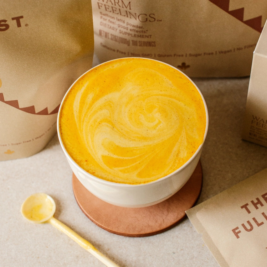 A close-up of a creamy, golden saffron latte with a swirled froth design on top, served in a white cup on a light brown leather coaster. A tiny spoon rests on the table in front.