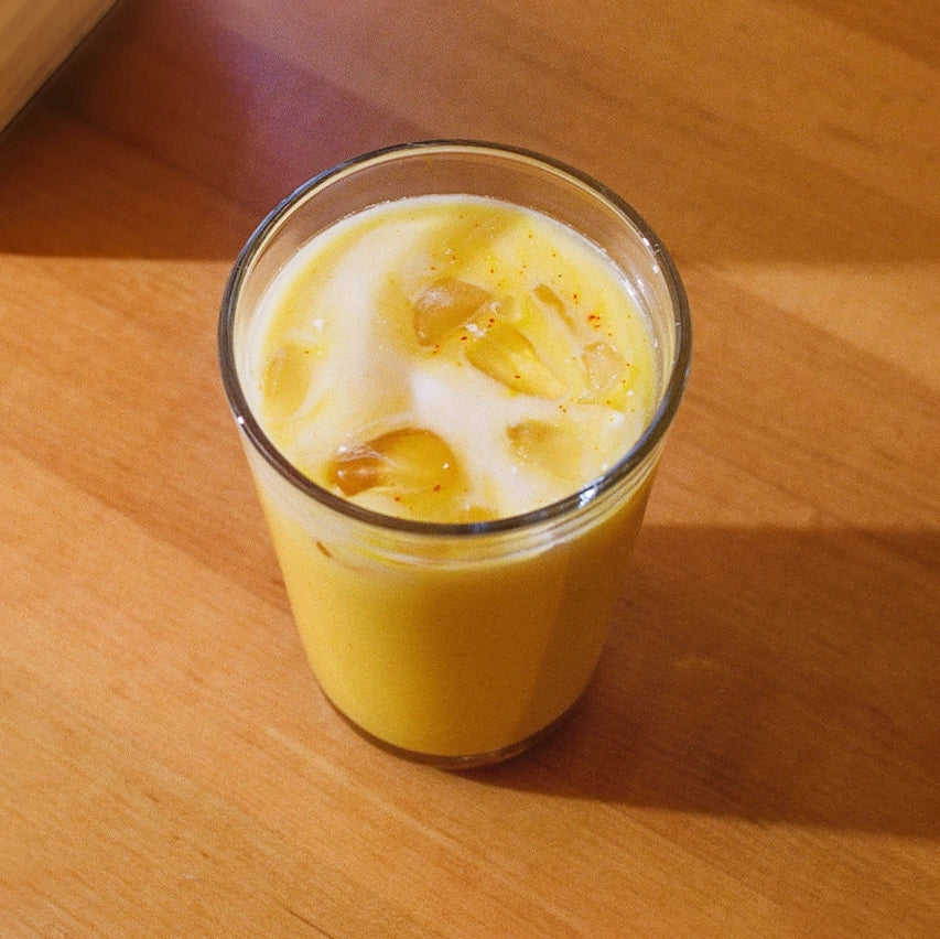A glass of chilled, yellow-orange saffron latte with ice cubes floating on top, casting a shadow on a wooden table under warm lighting, creating a refreshing visual.