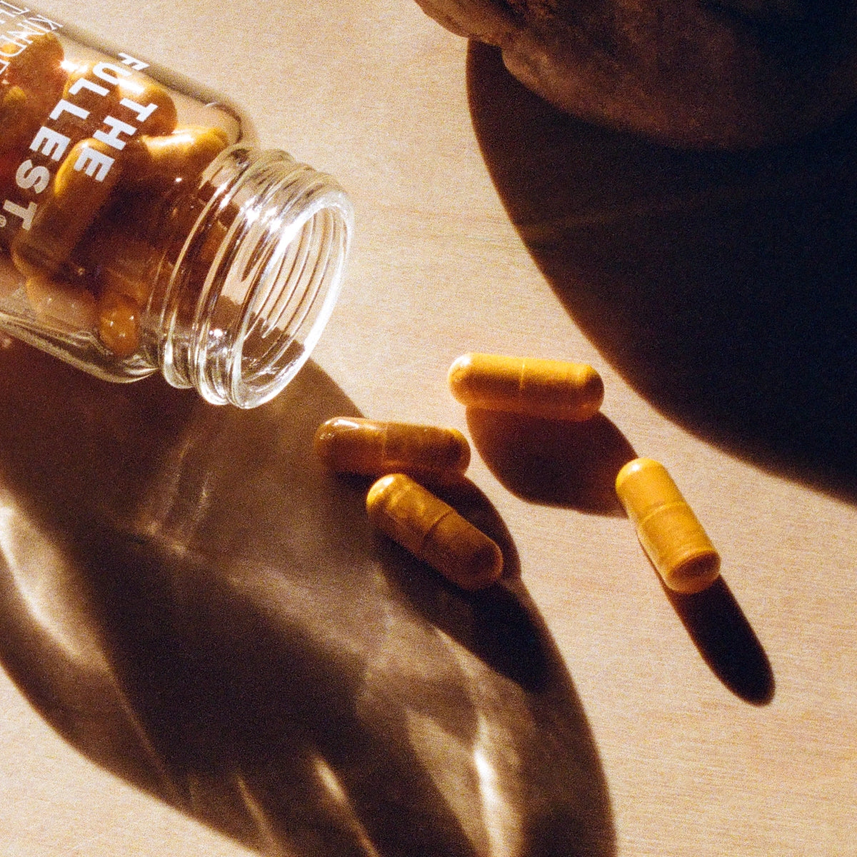 A clear bottle of THE FULLEST Saffron Capsules lies on its side, spilling several orange saffron capsules—onto a sunlit surface.