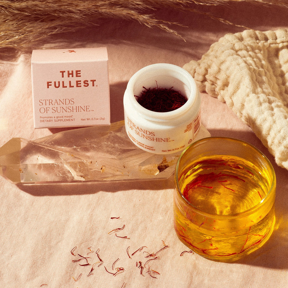 THE FULLEST &quot;Strands of Sunshine&quot;, made from pure saffron flower, is displayed with its packaging, alongside a clear cup of tea and scattered saffron threads on a textured surface.
