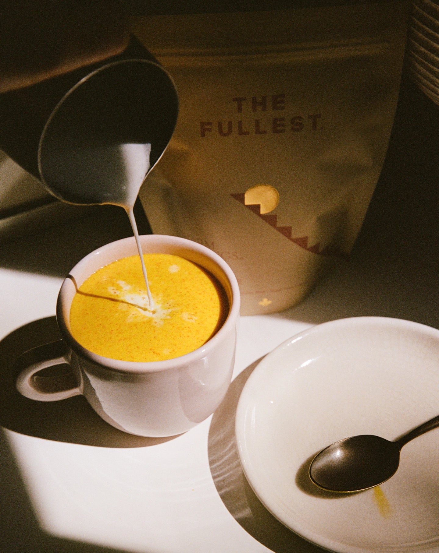 A white mug filled to the brim with a vibrant, saffron-colored frothy drink, as a stream of milk is being poured in, creating a delicate pattern on the surface. The mug sits next to a saucer.