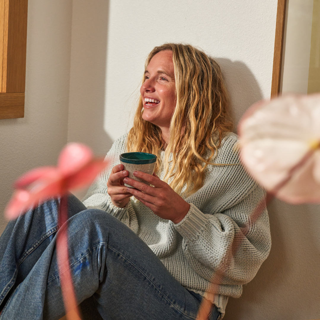A woman with long blonde hair sits on the floor, smiling and holding a mug of THE FULLEST saffron latte. She wears a light sweater and jeans, with out-of-focus pink flowers in the foreground. The scene radiates wellness.