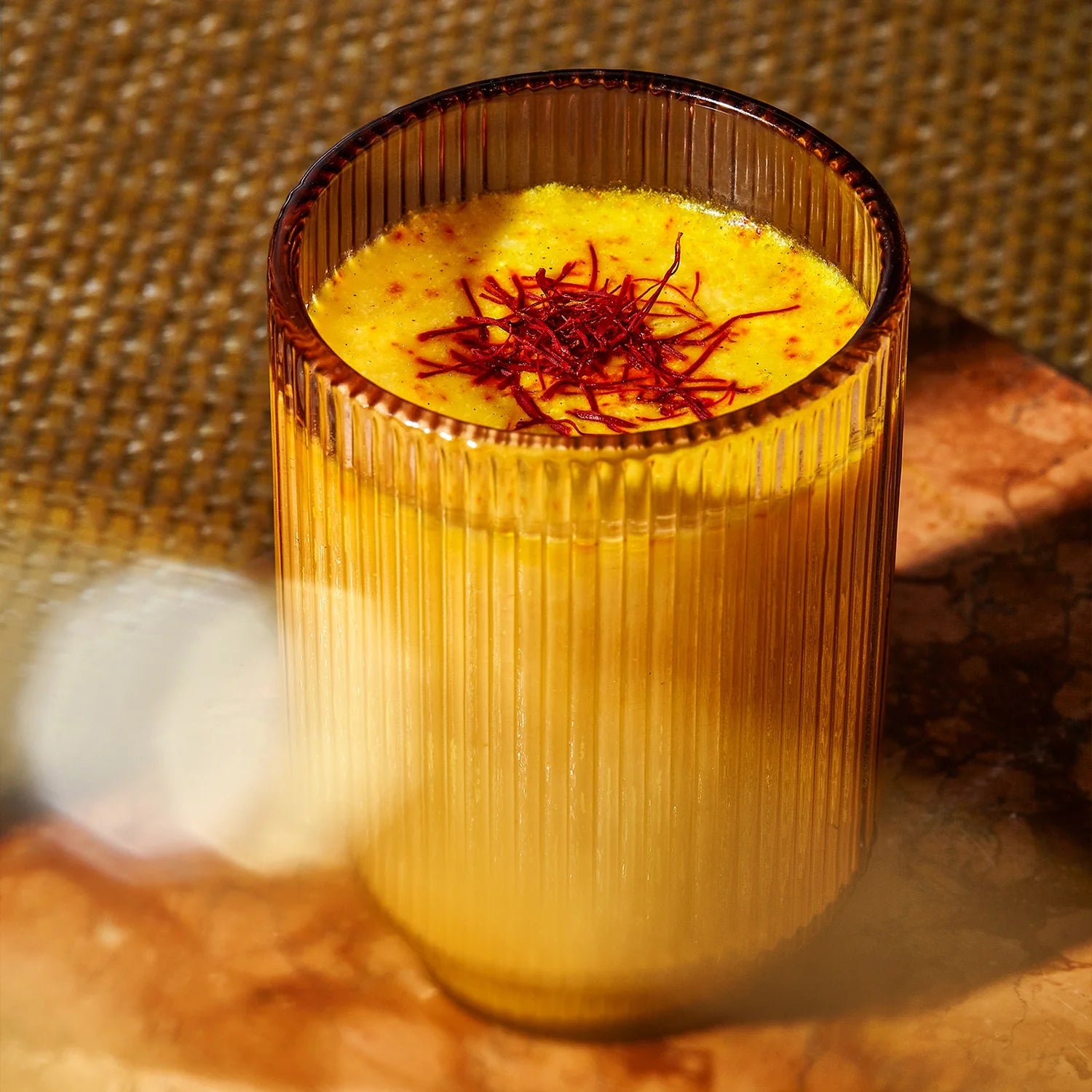 A glass of mango lassi topped with THE FULLEST Saffron Bundle sits on a brown marble surface with a woven mat in the background.