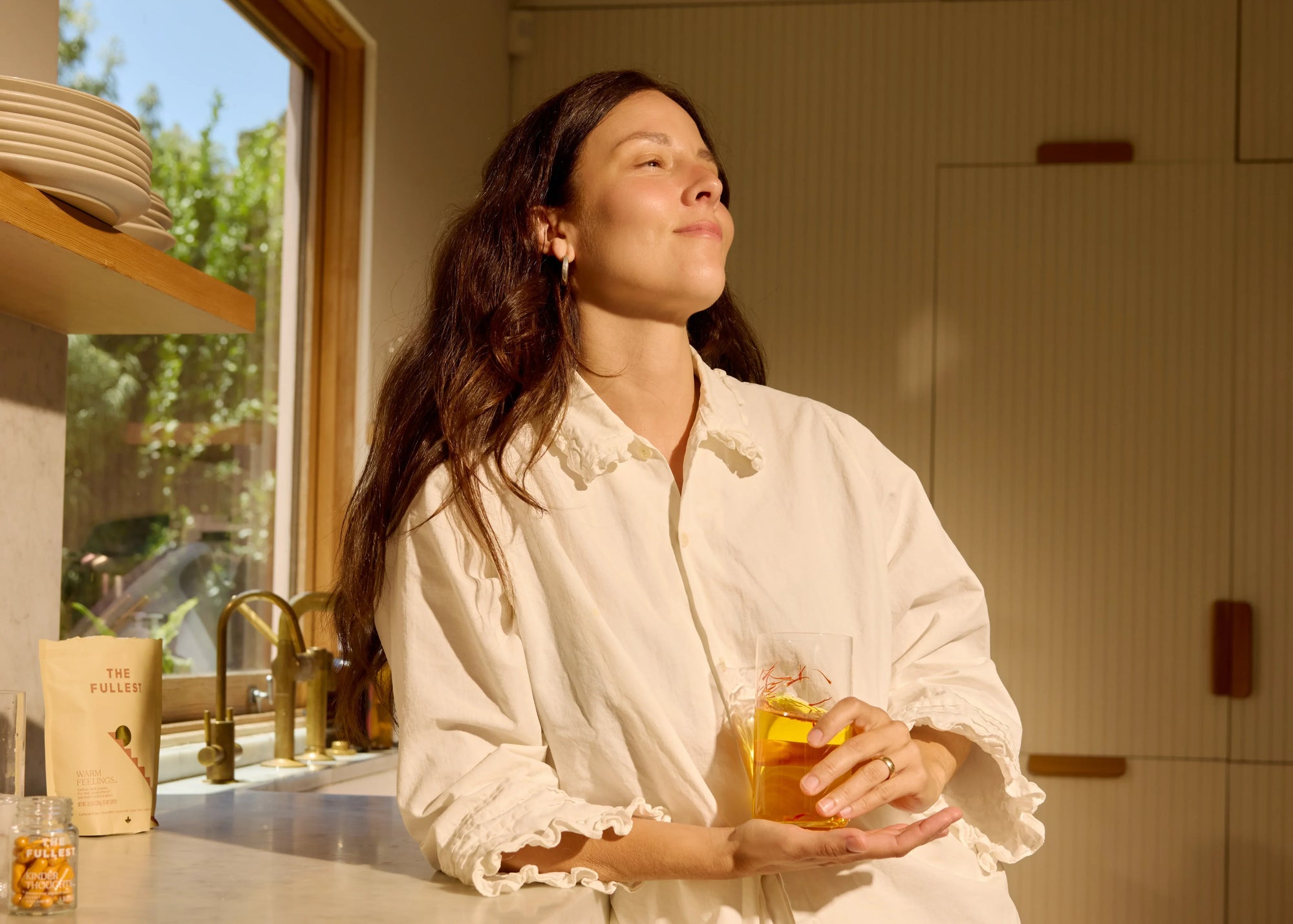Woman standing in natural lit kitchen with a glass of saffron water in hand 