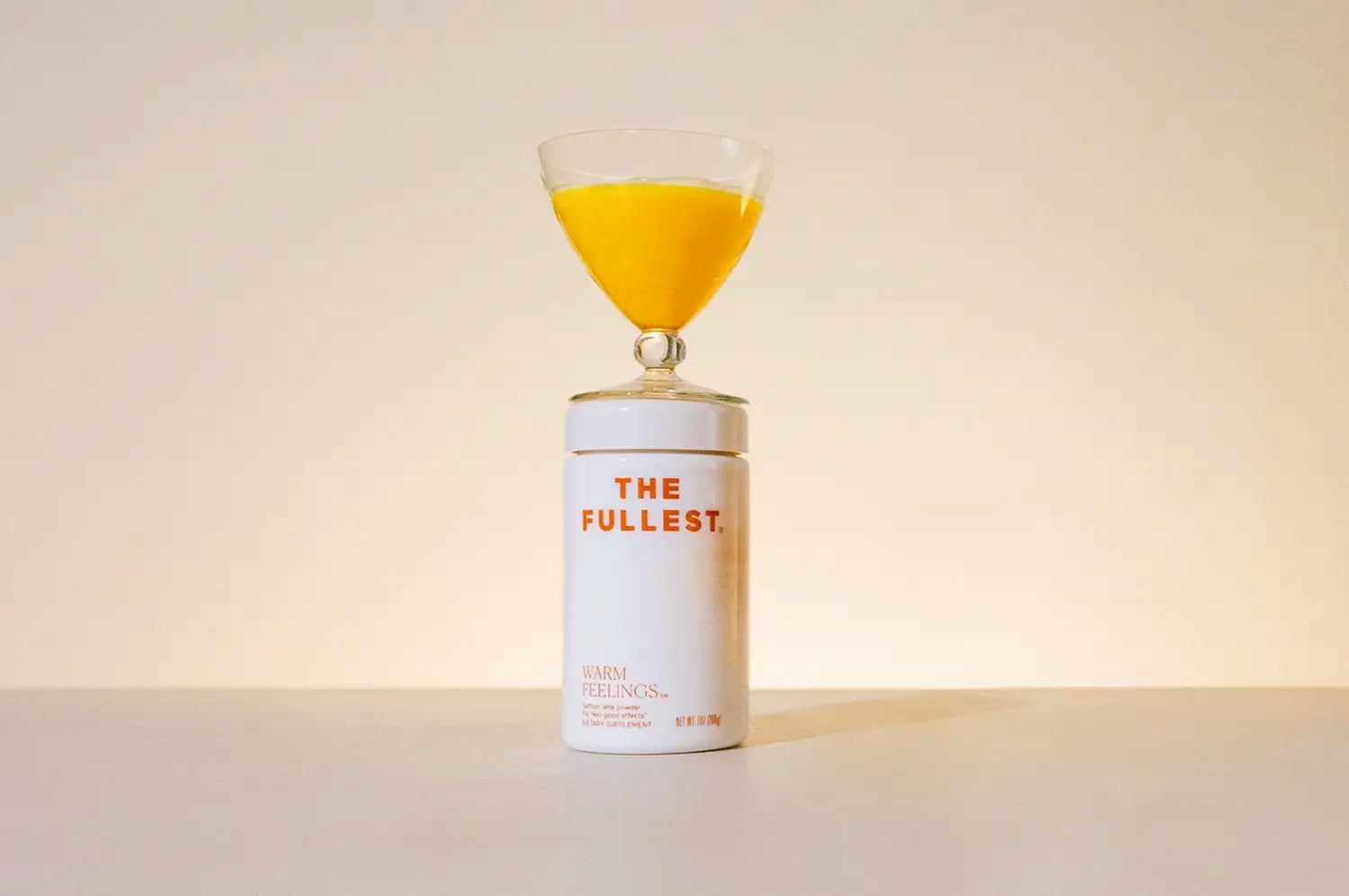 A glass of a saffron beverage balanced on top of it's white container labeled "the fullest" against a pale background.