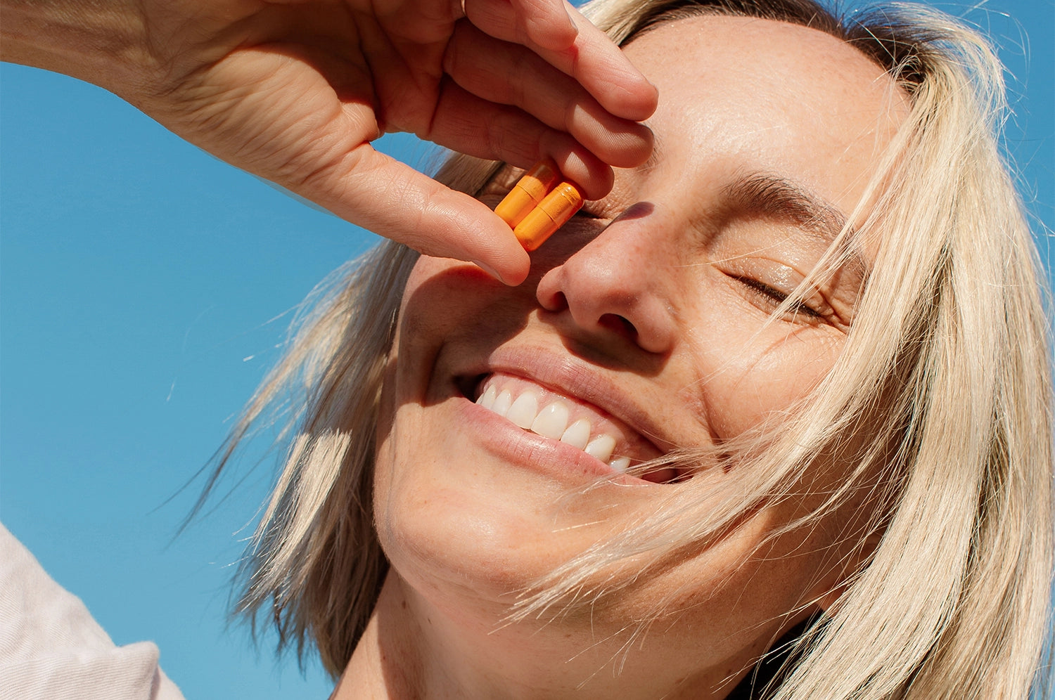 A smiling woman with short blonde hair holding two THE FULLEST Kinder Thoughts saffron and turmeric capsules against her face.