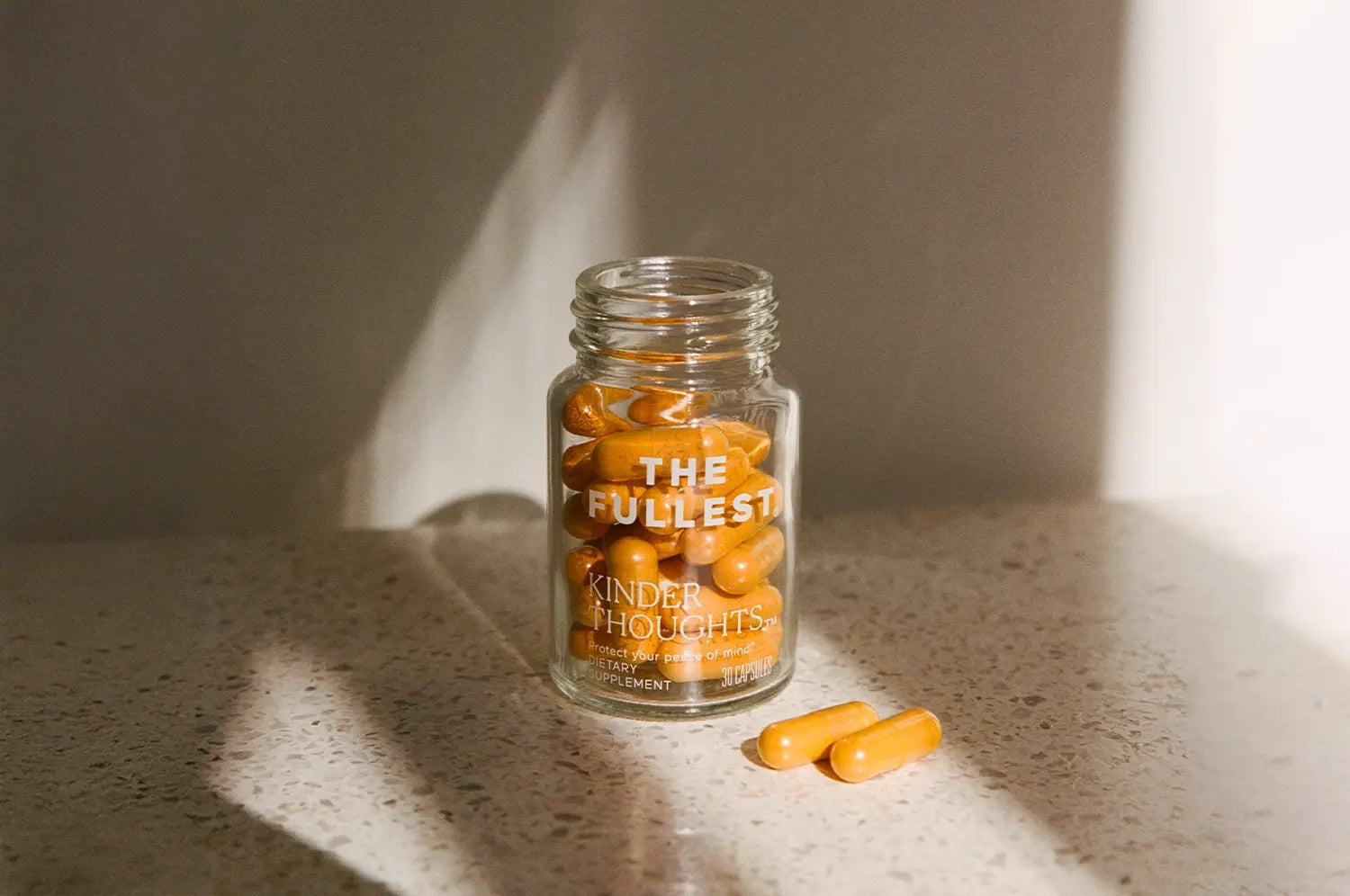 Bottle of THE FULLEST's Kinder Thoughts® saffron and turmeric capsules on a marble countertop with moody shadow.