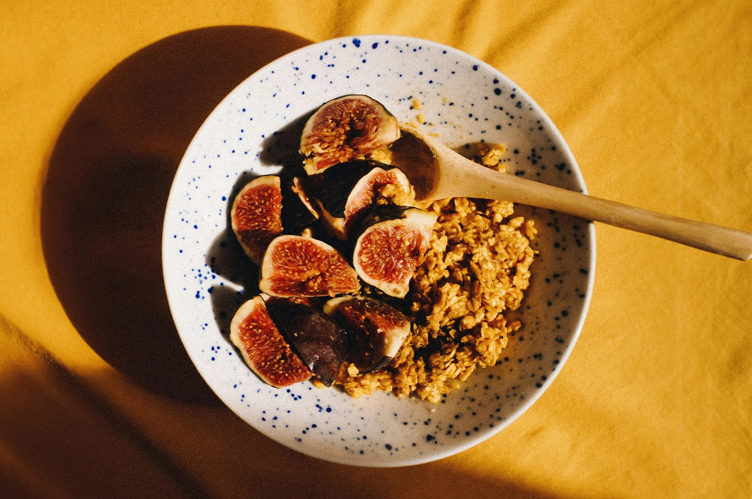 A bowl of saffron tahini granola with sliced fresh figs on top of a mustard colored table cloth.