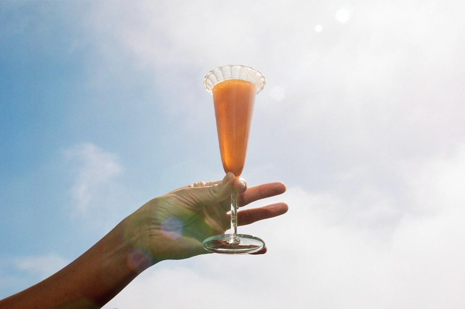 A hand elegantly holds a glass filled with a saffron spritz, its effervescent surface catching the sunlight against a bright blue sky. The glass, fluted with a scalloped rim, exudes a sense of calm and luxury, symbolizing a serene pre-bedtime ritual!