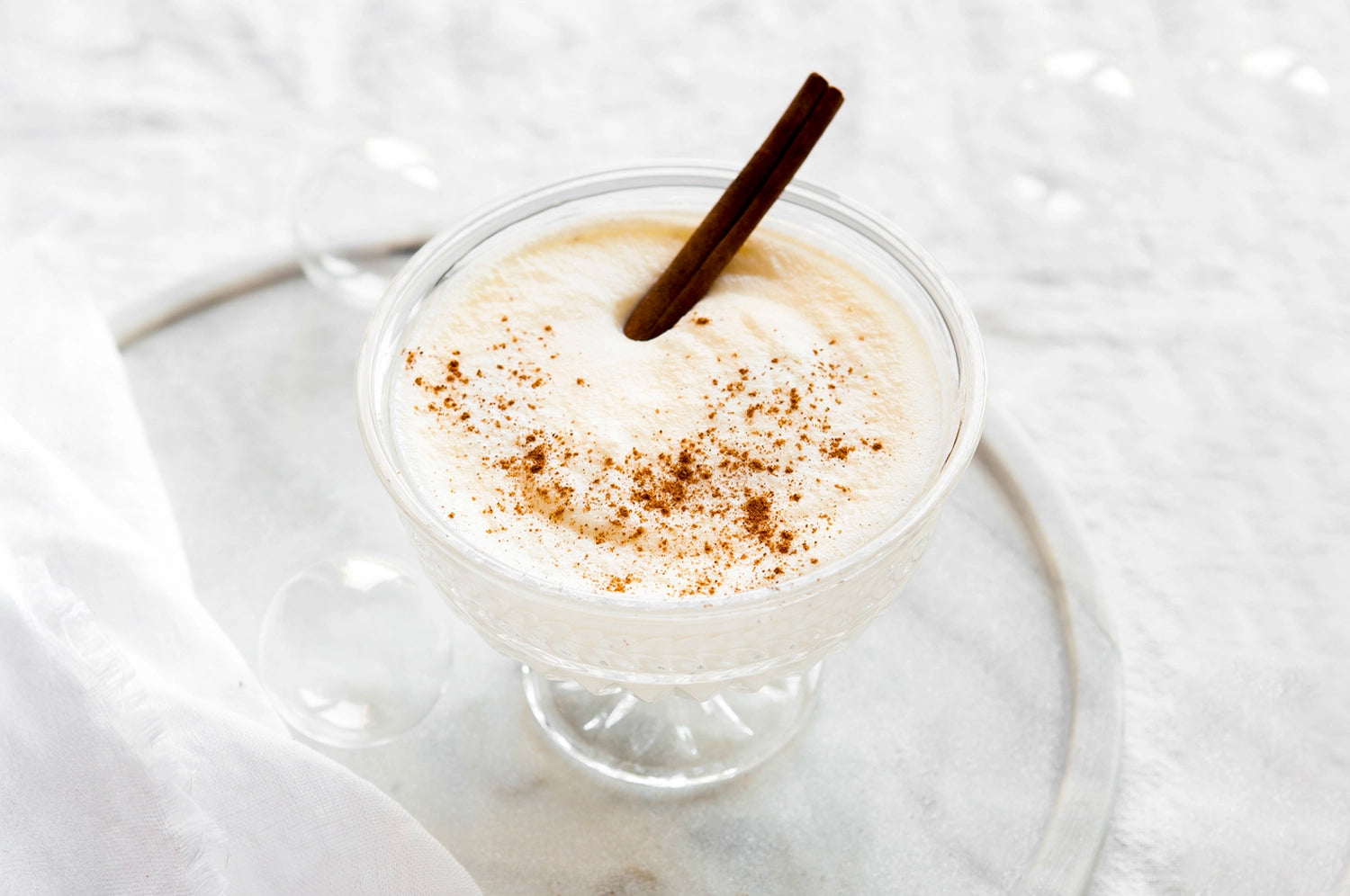 A creamy, frothy saffron eggnog served in a vintage-style glass with a dusting of nutmeg and a cinnamon stick garnish, sitting on a light marble surface with a soft, white cloth in the background.