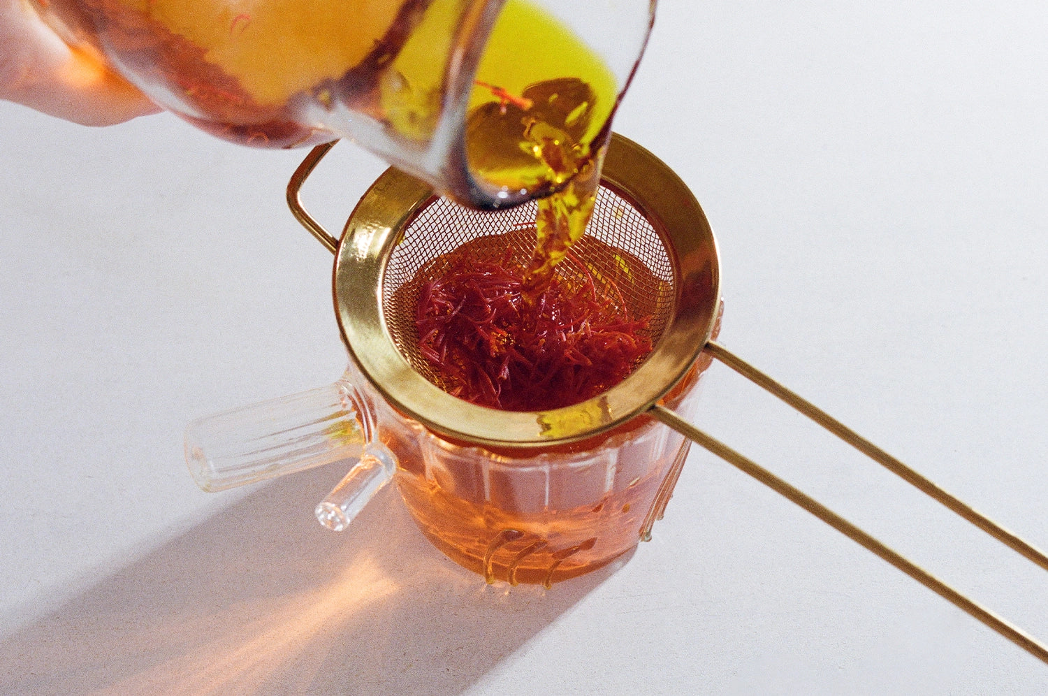 An overhead view of a model pouring The Fullest's saffron tea into a glass using a brass strainer.