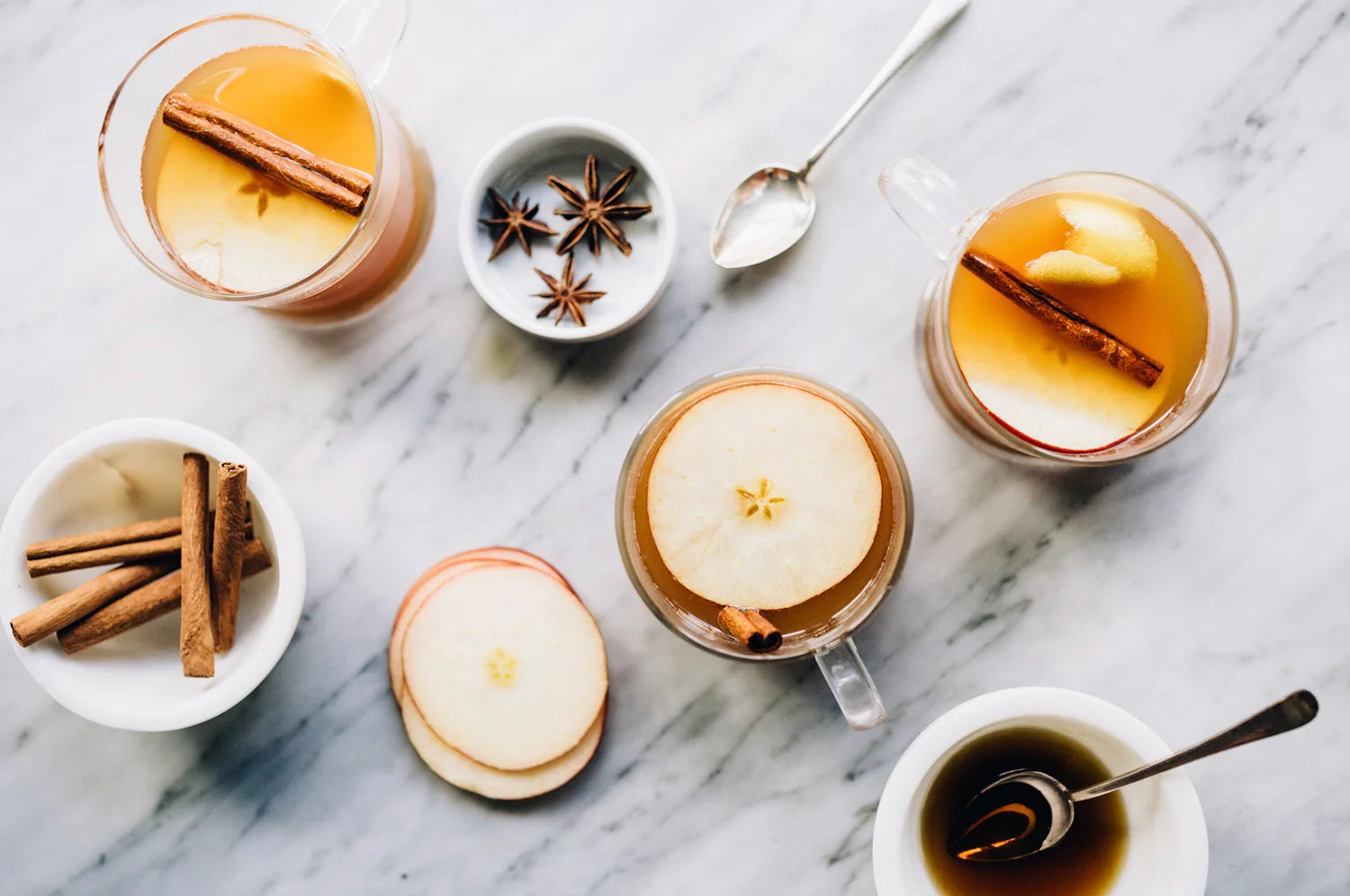 Overhead view of saffron mulled apple cider in glass mugs, garnished with apple slices, cinnamon sticks, and lemon peel. Surrounding the drinks are star anise, cinnamon sticks, sliced apples, and a small bowl of tea, all arranged on a marble surface.