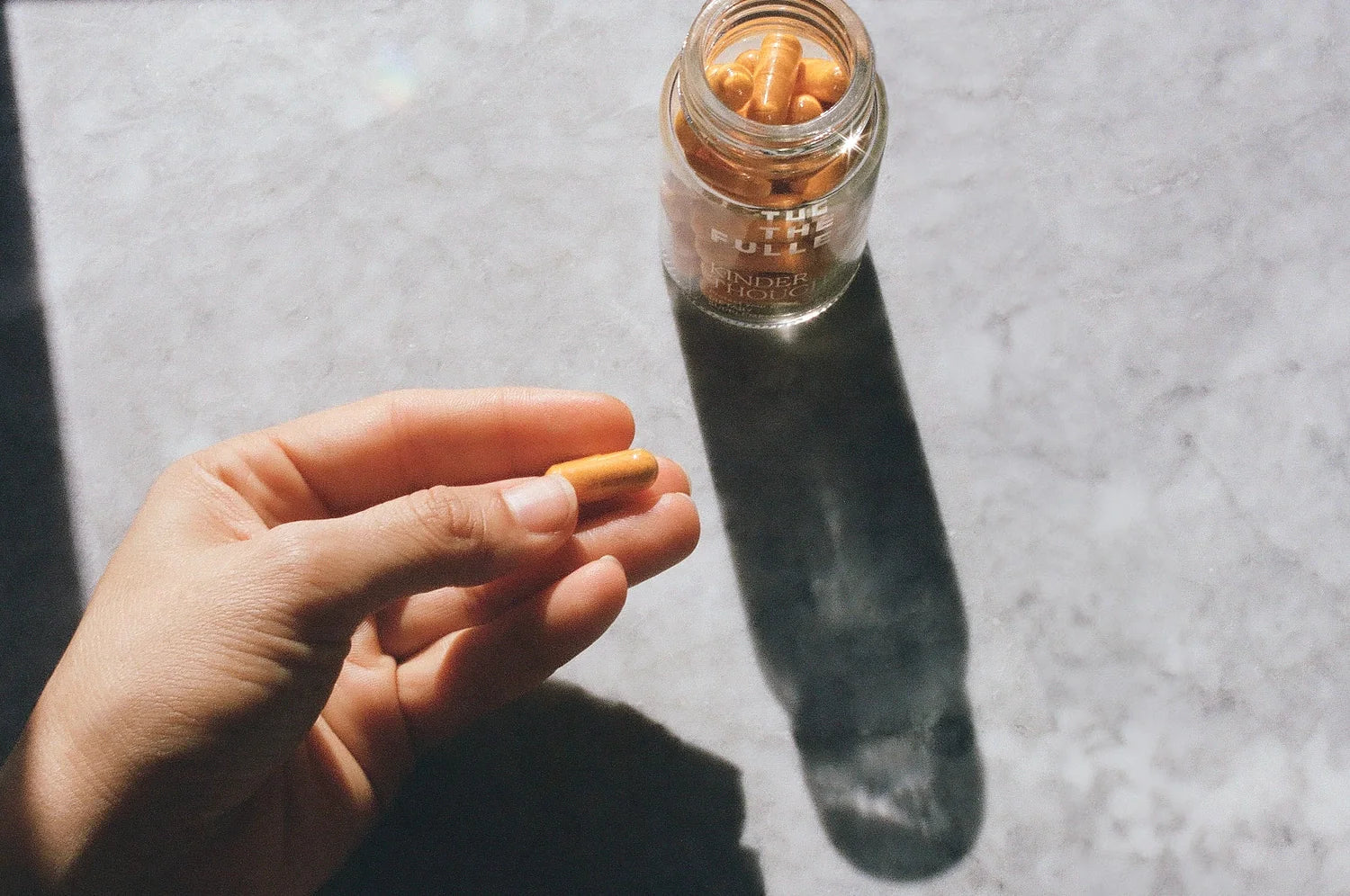 An overhead photo showing a woman holding a Kinder Thoughts capsule, with the clear pill bottle labeled "The Fullest Kinder Thoughts" filled with orange capsules, placed on the counter.