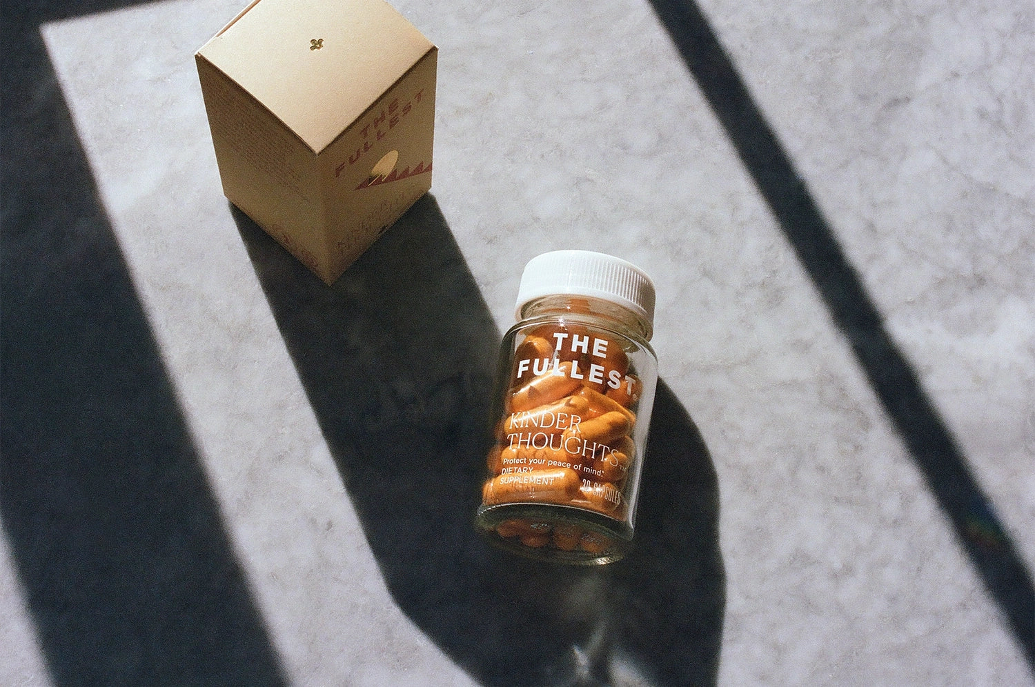 An overhead view of a clear pill bottle labeled "the fullest kinder thoughts," filled with orange saffron and turmeric capsules, placed flat on a grey concrete surface next to its box.