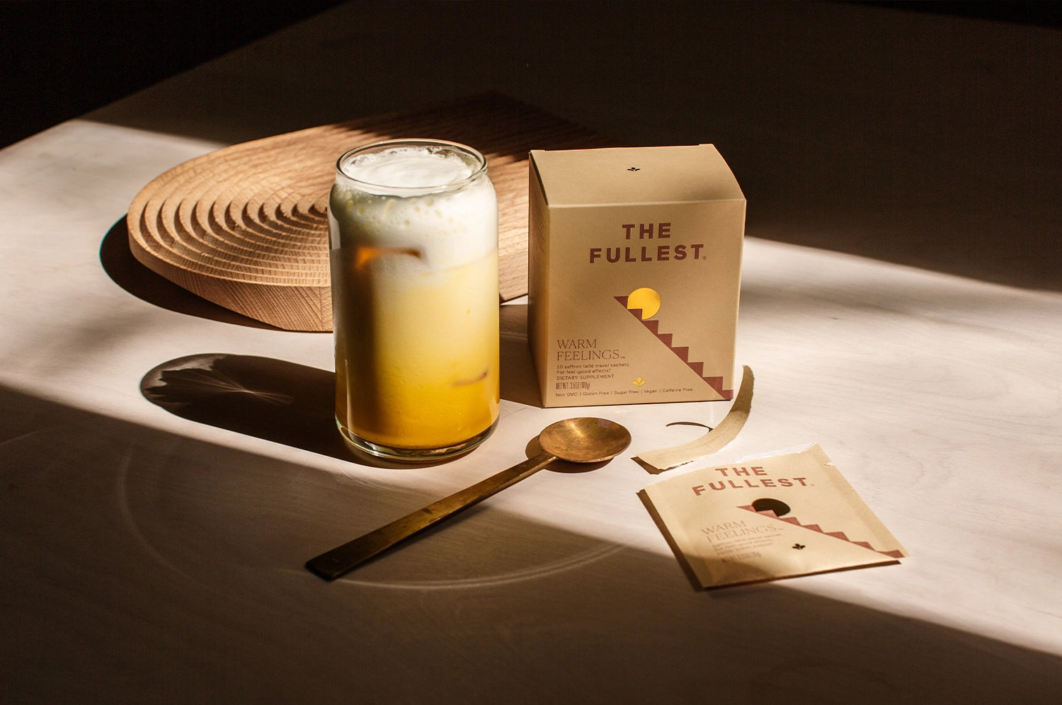 THE FULLEST Warm Feelings saffron latte drink in glass cup sitting next to brass spoon and warm feelings sachet box.
