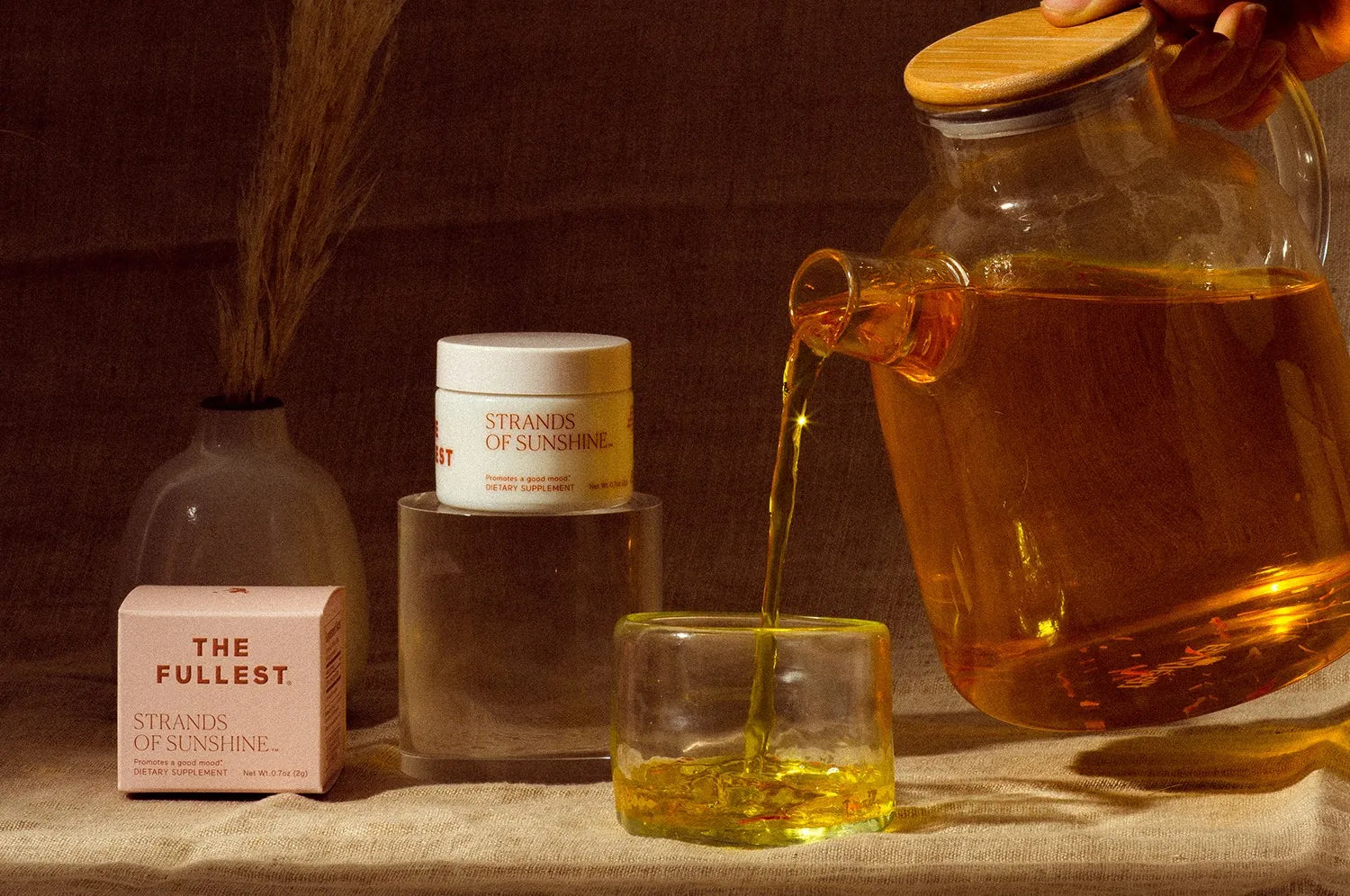 A frontal image of a model holding a clear teapot filled with Saffron tea from The Fullest. The Strands of Sunshine saffron threads are in their white container, placed outside next to the branded packaging box. 