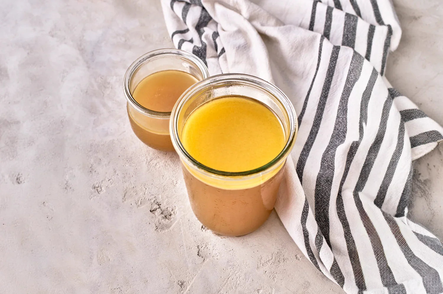 THE FULLEST saffron bone broth in two mason jars next to a striped towel on a concrete surface.