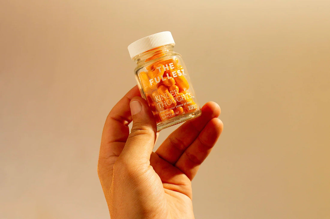 A hand holding a bottle of THE FULLEST's Kinder Thoughts saffron capsules against a tan background.