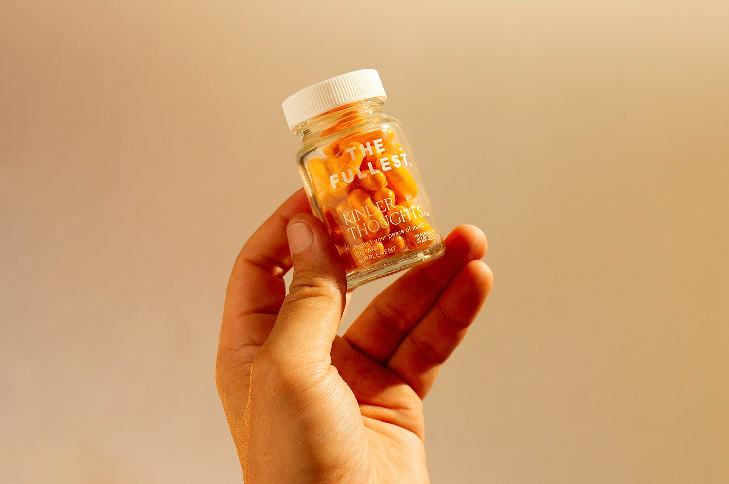 A hand holding a bottle of THE FULLEST's Kinder Thoughts saffron capsules against a tan background.