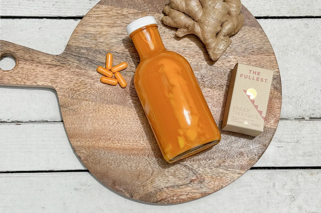A bottle of THE FULLEST's Kinder Thoughts saffron drinking vinegar, Kinder Thoughts saffron capsules, and fresh ginger sitting on a wood cutting board that rests atop a paneled wood surface.