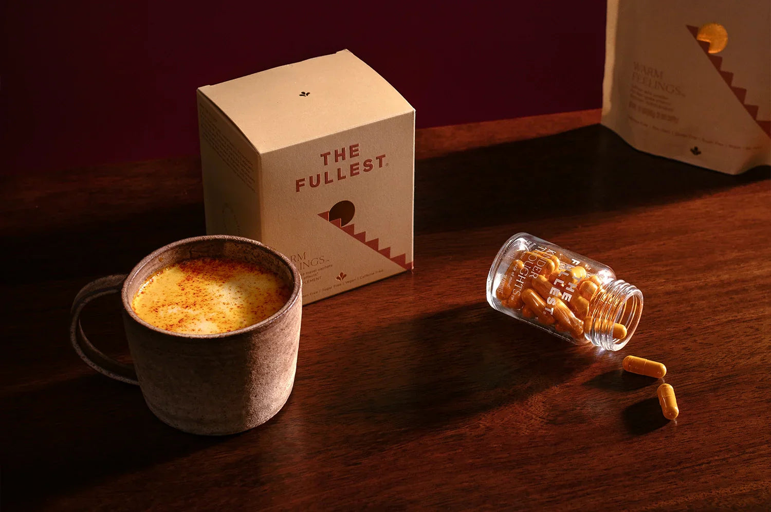 A cozy scene featuring a warm saffron latte in a rustic ceramic mug, a box of THE FULLEST’s Warm Feelings saffron latte mix, and a glass jar of Kinder Thoughts saffron capsules on a wooden surface.