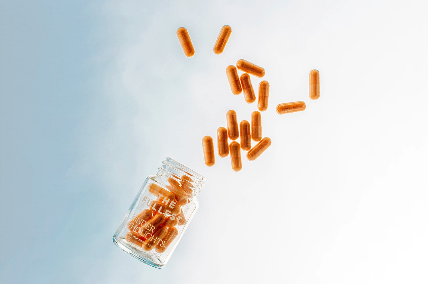 Saffron and turmeric Kinder Thoughts capsules from The Fullest spill out of a glass bottle against a bright blue sky, forming an arc above the bottle.