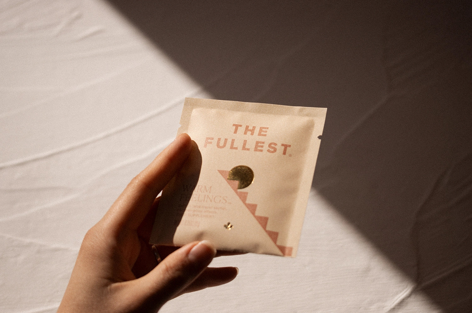 A hand holding a Warm Feelings saffron latte sachet from THE FULLEST in direct sunlight against a white wall.