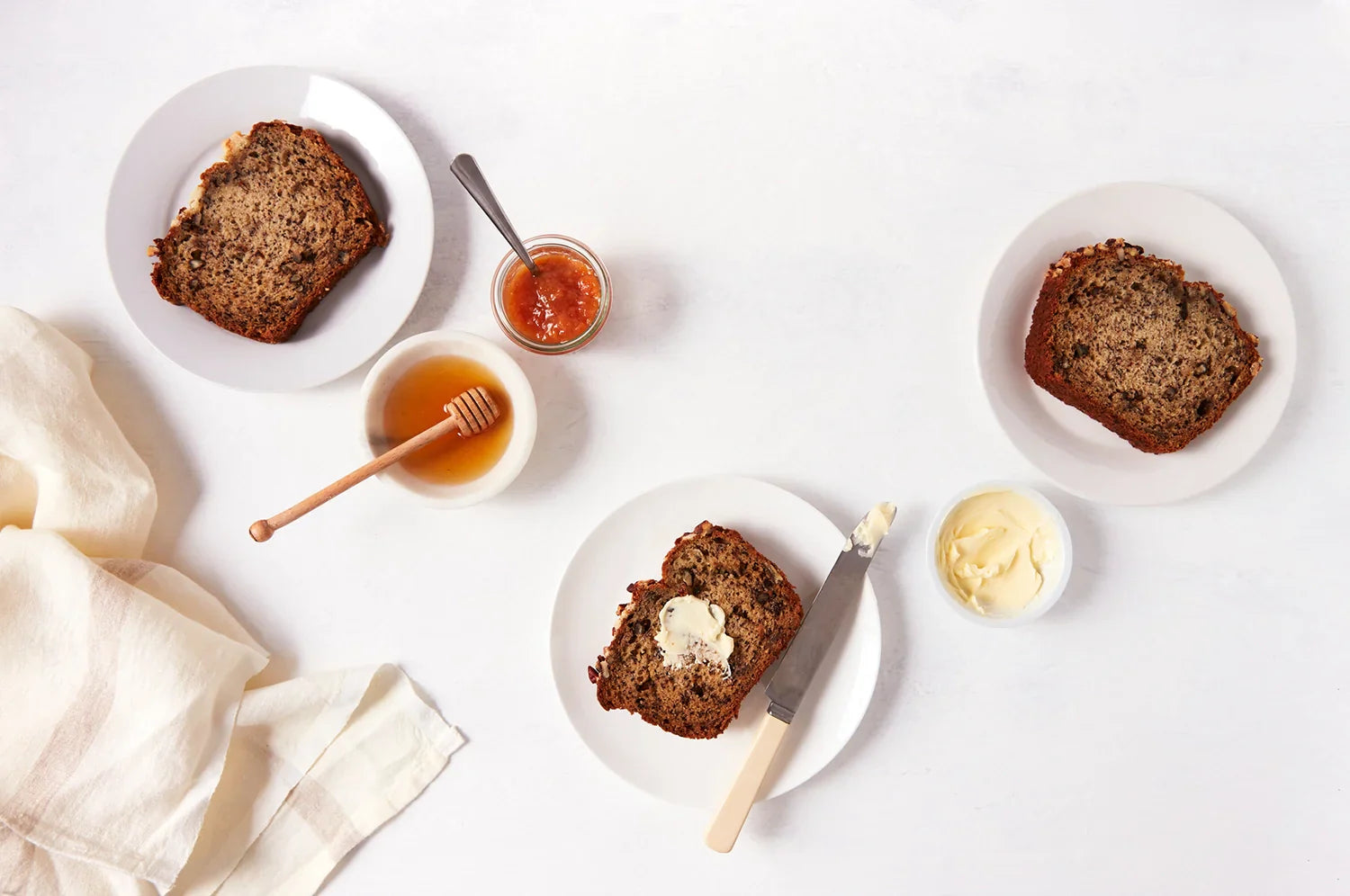 Slices of THE FULLEST saffron banana bread on white plates with small bowls of honey, preserves, and butter next to them.