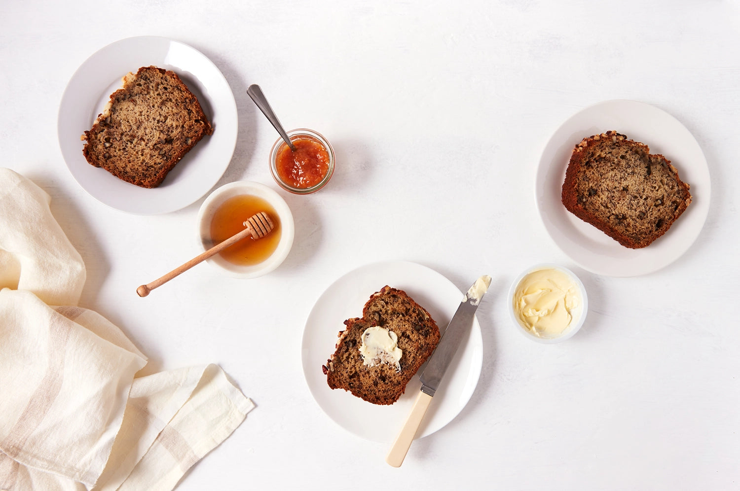 Slices of THE FULLEST's Warm Feelings saffron banana bread on white plates with small bowls of honey, preserves, and butter next to them.