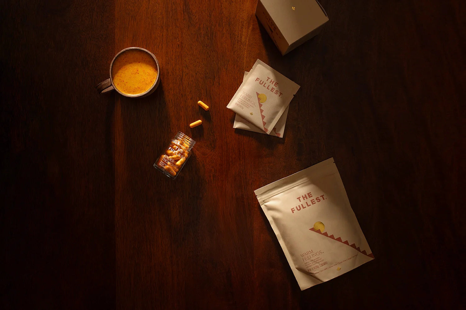 THE FULLEST products on a dark wooden table, featuring a saffron latte, Kinder Thoughts capsules, and Warm Feelings latte packets and pouch, warmly lit to showcase their premium packaging.