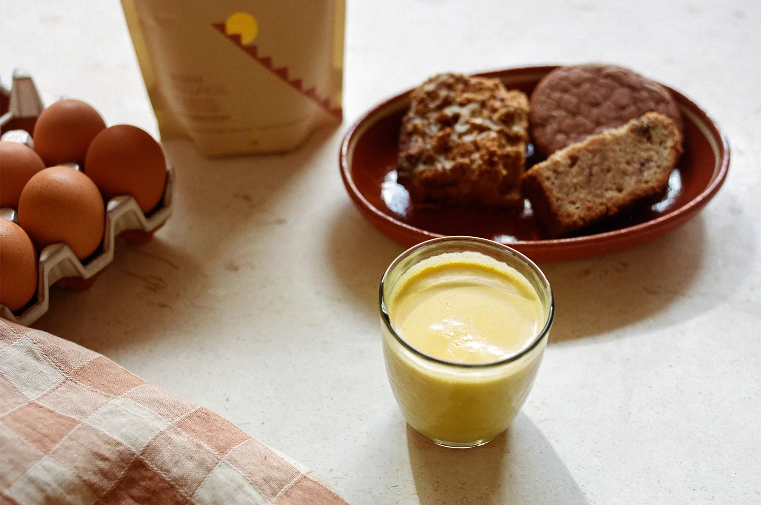 A cozy kitchen scene featuring a glass of golden saffron latte on a light countertop. Nearby, there is a plate with slices of banana bread and a cookie, a carton of brown eggs, and a beige and terracotta-colored cloth with a checkered pattern.