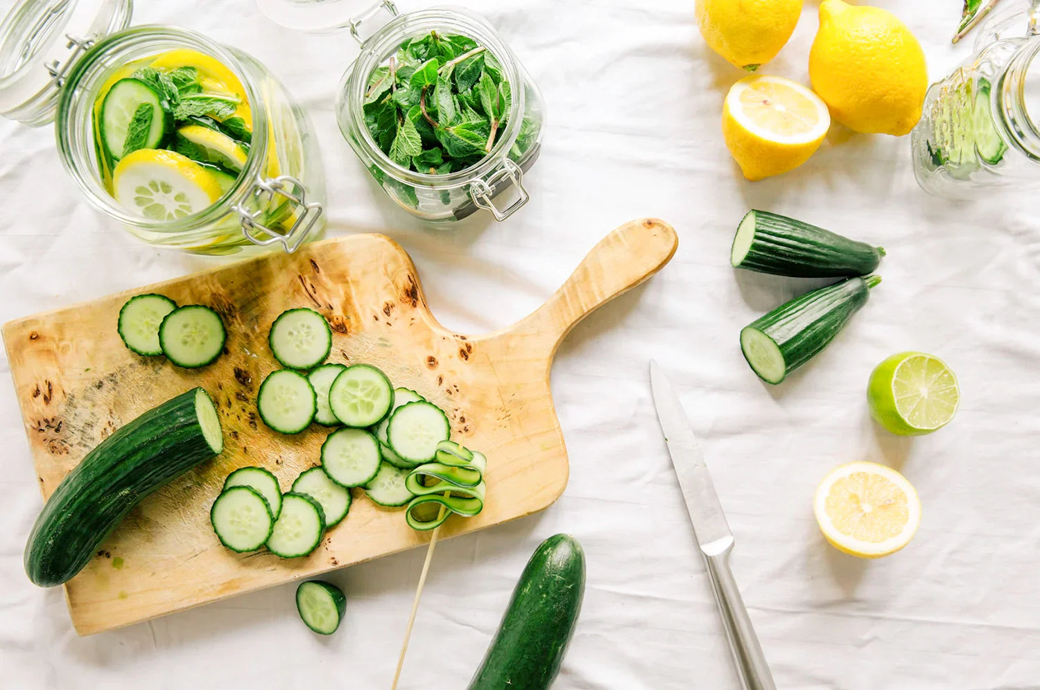 An overhead photo looking down on a scene of cucumber salad prep: a cutting board with a cucumber atop it being sliced, lemons sliced alongside it, and fresh mint in a jar.