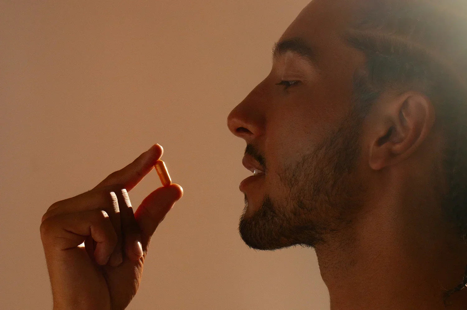 Profile view of a man gently holding a Kinder Thoughts capsule between his thumb and forefinger, set against a minimalist tan background, highlighting the elegance and simplicity of THE FULLEST’s saffron and turmeric supplement.