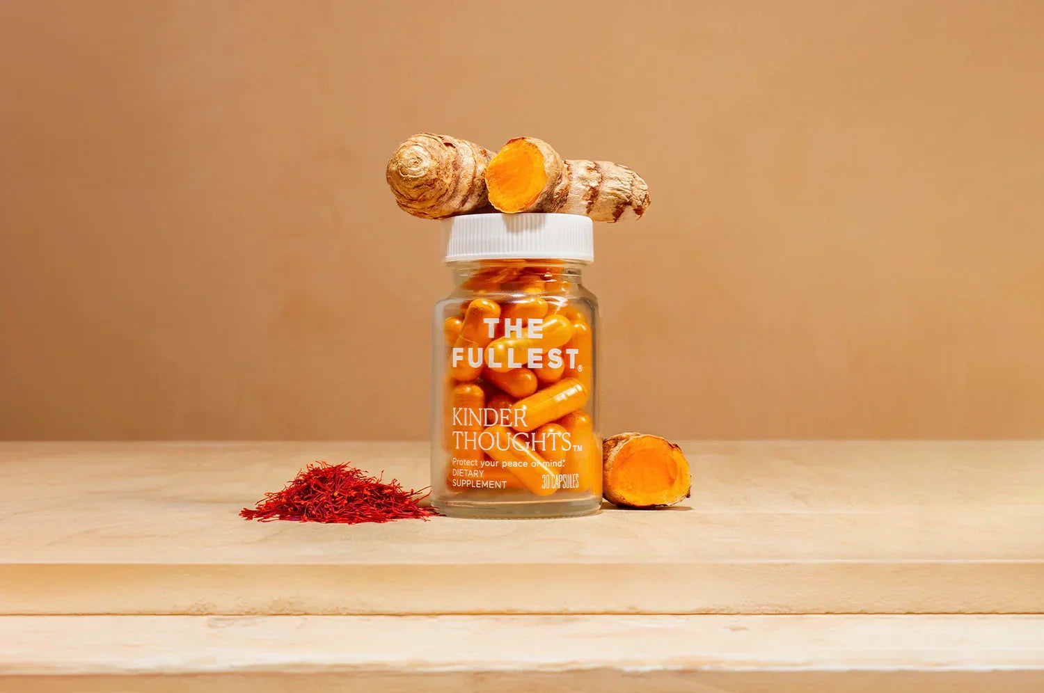 A bottle of THE FULLEST Saffron Capsules on a wood surface with fresh turmeric resting atop the botle and next to it, as well as a pile of saffron threads to the left of it.