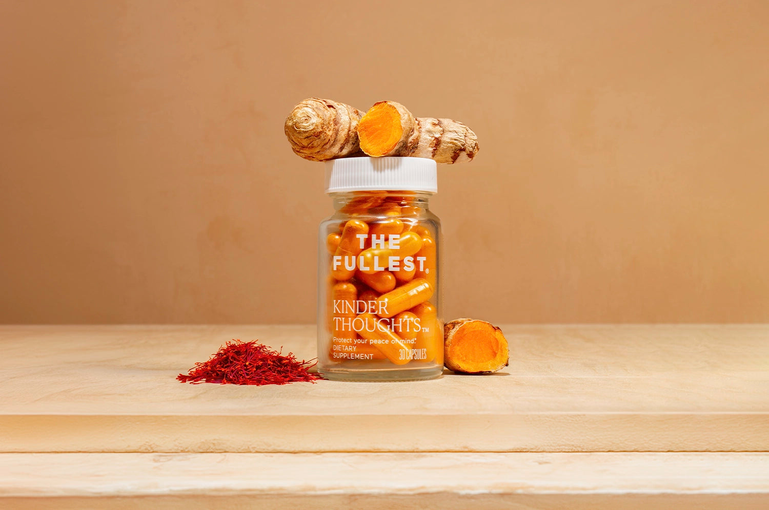 A bottle of THE FULLEST's Kinder Thoughts saffron capsules on a wood surface with fresh turmeric resting atop the botle and next to it, as well as a pile of saffron threads to the left of it.