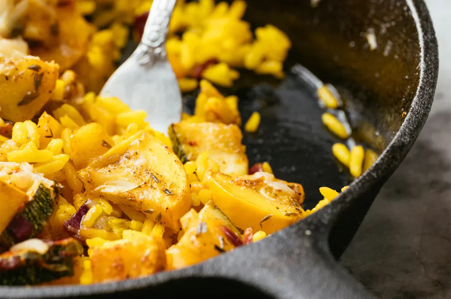 A cast iron skillet with THE FULLEST's saffron-infused risotto, roasted zucchini, and melted Parmesan. A silver fork rests in the creamy, herb-speckled dish, highlighting its warm, rustic appeal.
