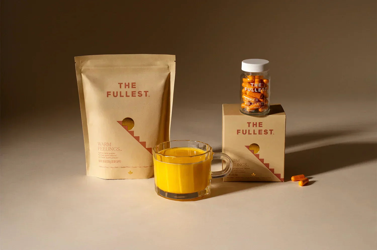 THE FULLEST products displayed in warm lighting: a kraft pouch of “Warm Feelings” saffron latte blend, a glass mug of golden latte, and a bottle of “Kinder Thoughts” saffron capsules on a matching box with two capsules in the foreground.