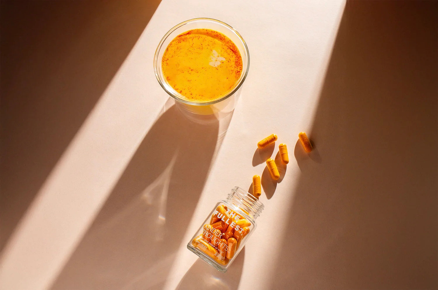 This image showcases a glass of golden-hued saffron latte and a scattered handful of orange capsules next to an open glass jar labeled “THE FULLEST Kinder Thoughts.”