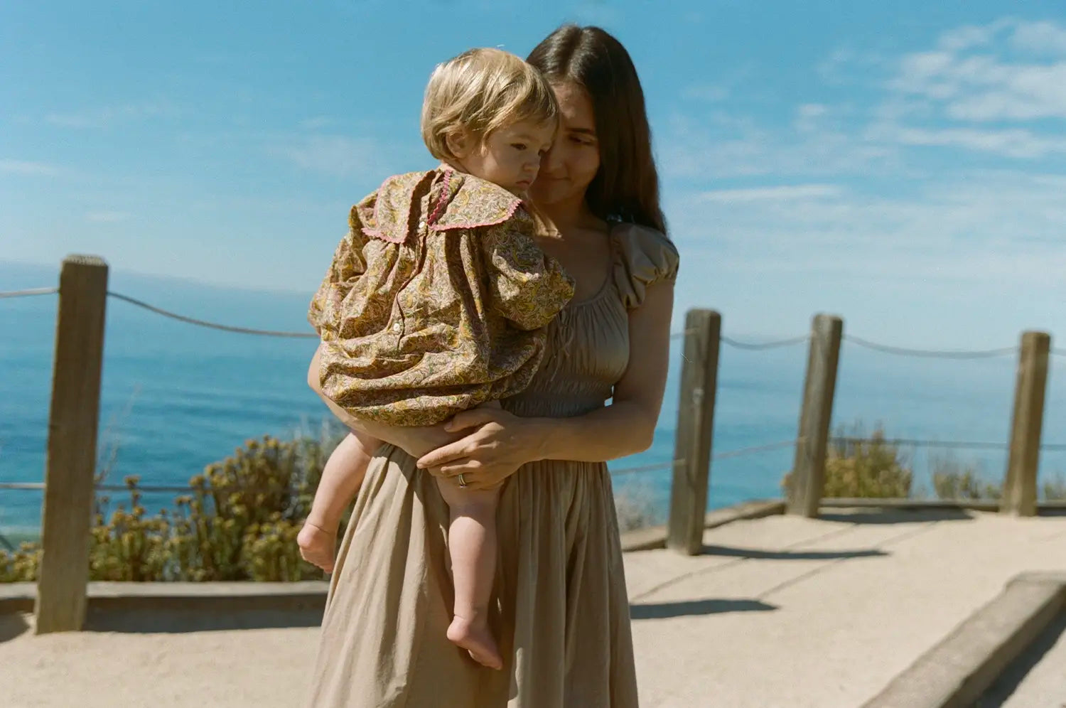 Mother holding daughter on a walkway with the ocean in the background 