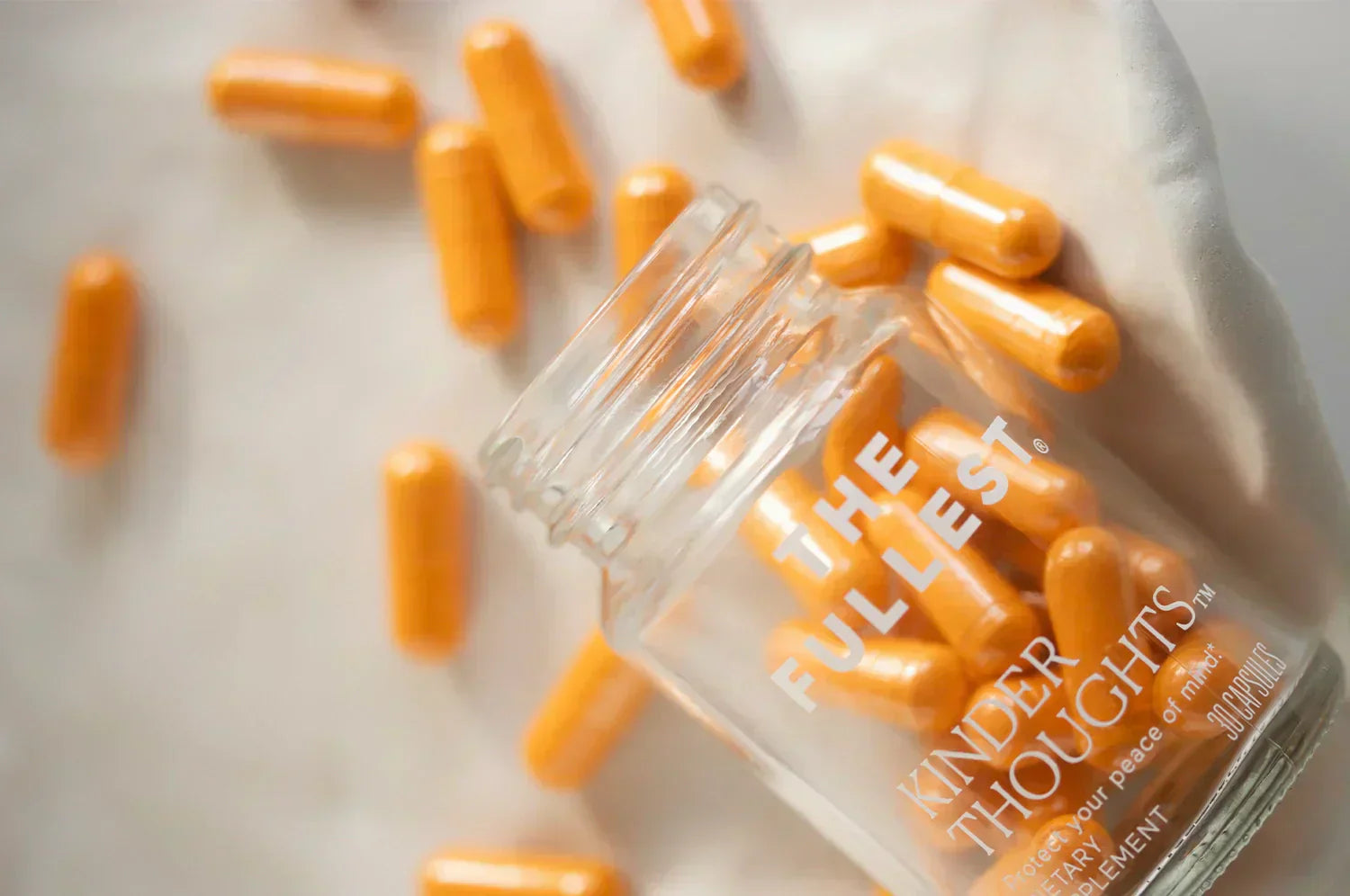 Glass jar of THE FULLEST Kinder Thoughts® saffron capsules spilling onto a soft fabric surface, showcasing vibrant orange capsules designed to support mood and mental well-being.