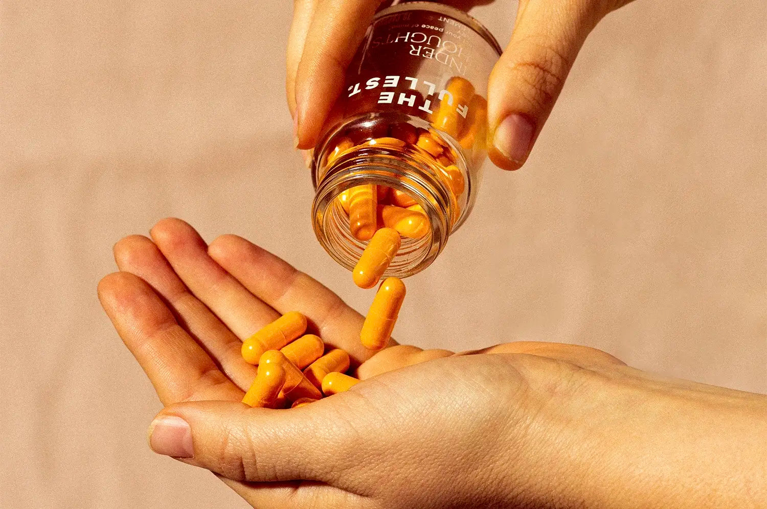 Close-up of hands pouring THE FULLEST's Kinder Thoughts capsules into an open palm. The capsules catch the light, emphasizing their glossy texture. The background is a neutral beige tone, complementing the warm color palette of the scene.