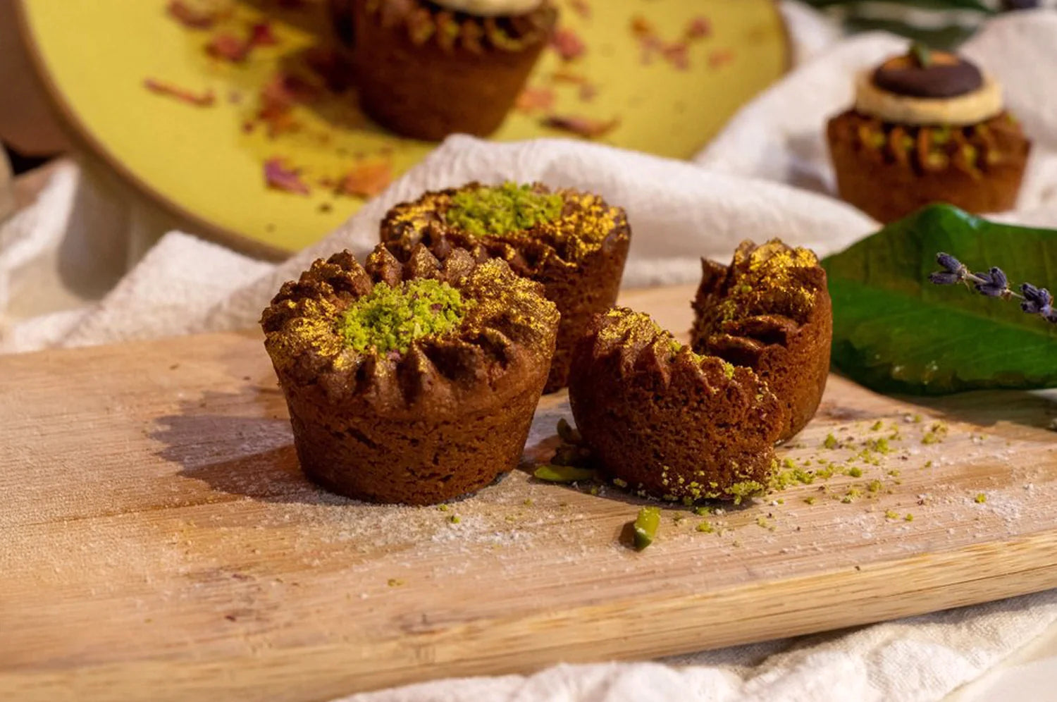 Golden-brown grain-free muffins topped with a dusting of gold powder and crushed pistachios, arranged on a rustic wooden board. The muffins have a crinkled, textured top, creating a visually appealing contrast with the vibrant green pistachio garnish.