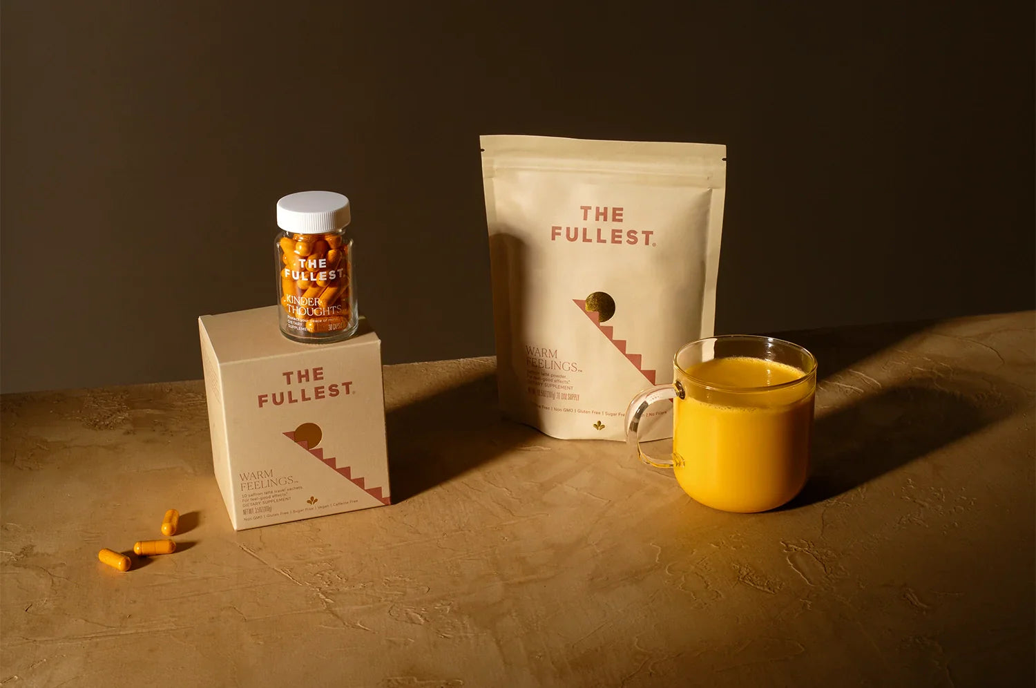 THE FULLEST saffron-based products displayed on a textured surface. Includes a jar of ‘Kinder Thoughts’ capsules on a ‘Warm Feelings’ box, a pouch labeled ‘Warm Feelings,’ and a glass mug with a Warm Feelings saffron latte prepared.
