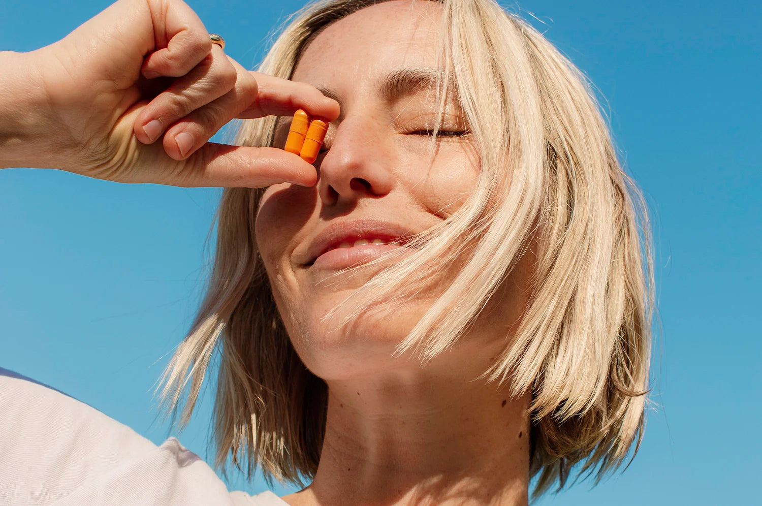 A woman with short blonde hair standing against a blue sky while holding two of THE FULLEST saffron capsules against her face.