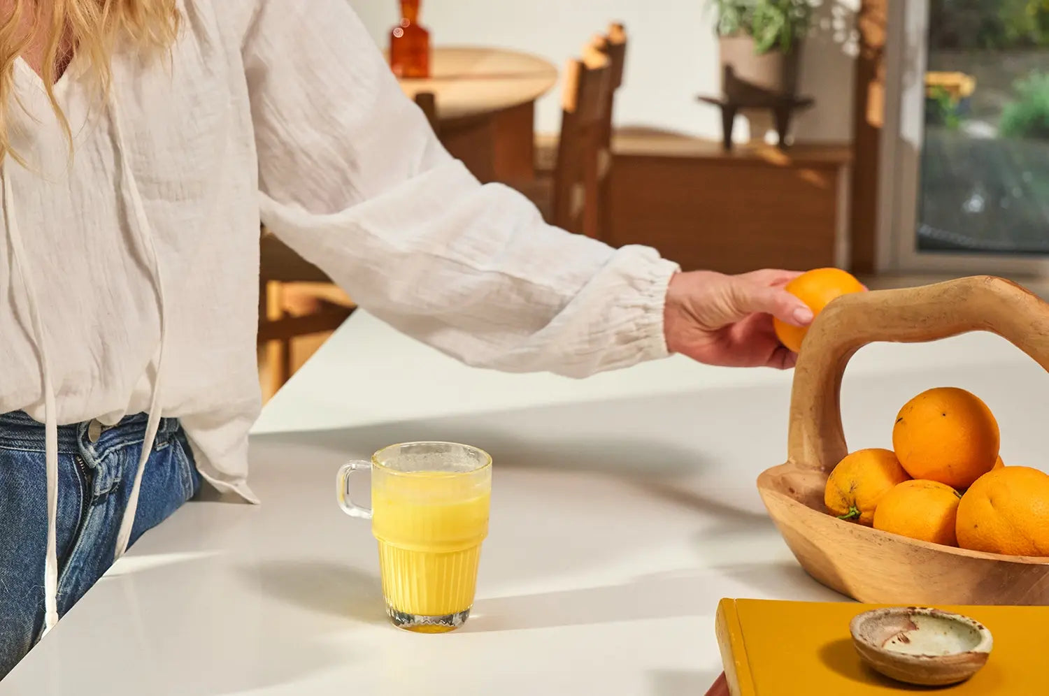 Woman reaching for an orange in a bright kitchen with a saffron latte placed nearby, capturing a moment of mindful morning routine and wellness.